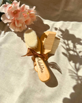 A personalized wooden baby brush and comb set engraved with the name "Adeline," tied together with a brown ribbon and softly lit by sunlight. The set is placed on a neutral fabric surface with soft shadows and pink floral accents.