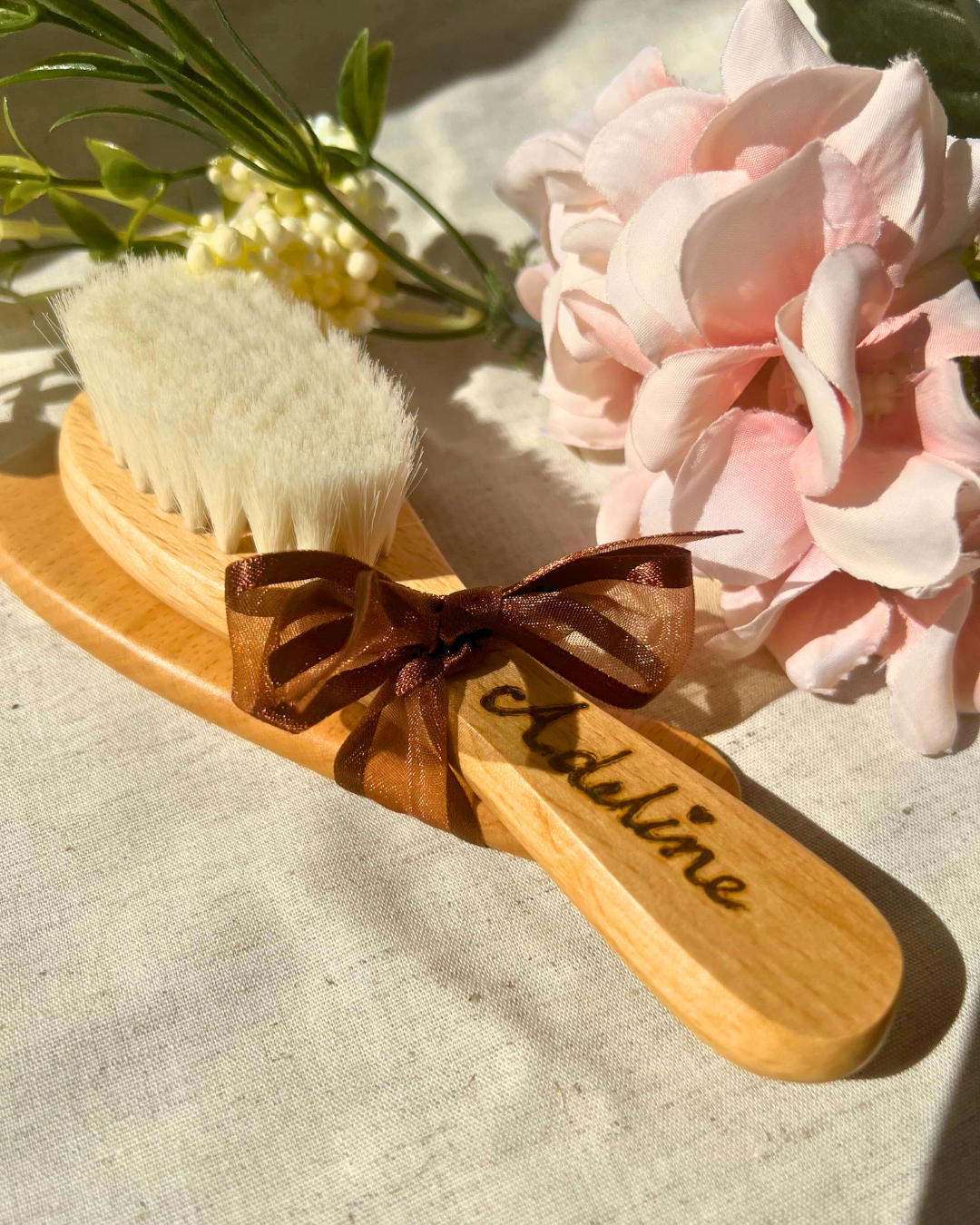 A soft-bristled wooden baby hairbrush engraved with the name "Adeline," tied with a brown organza ribbon and styled with gentle lighting, delicate pink flowers, and creamy white blossoms on a linen background.