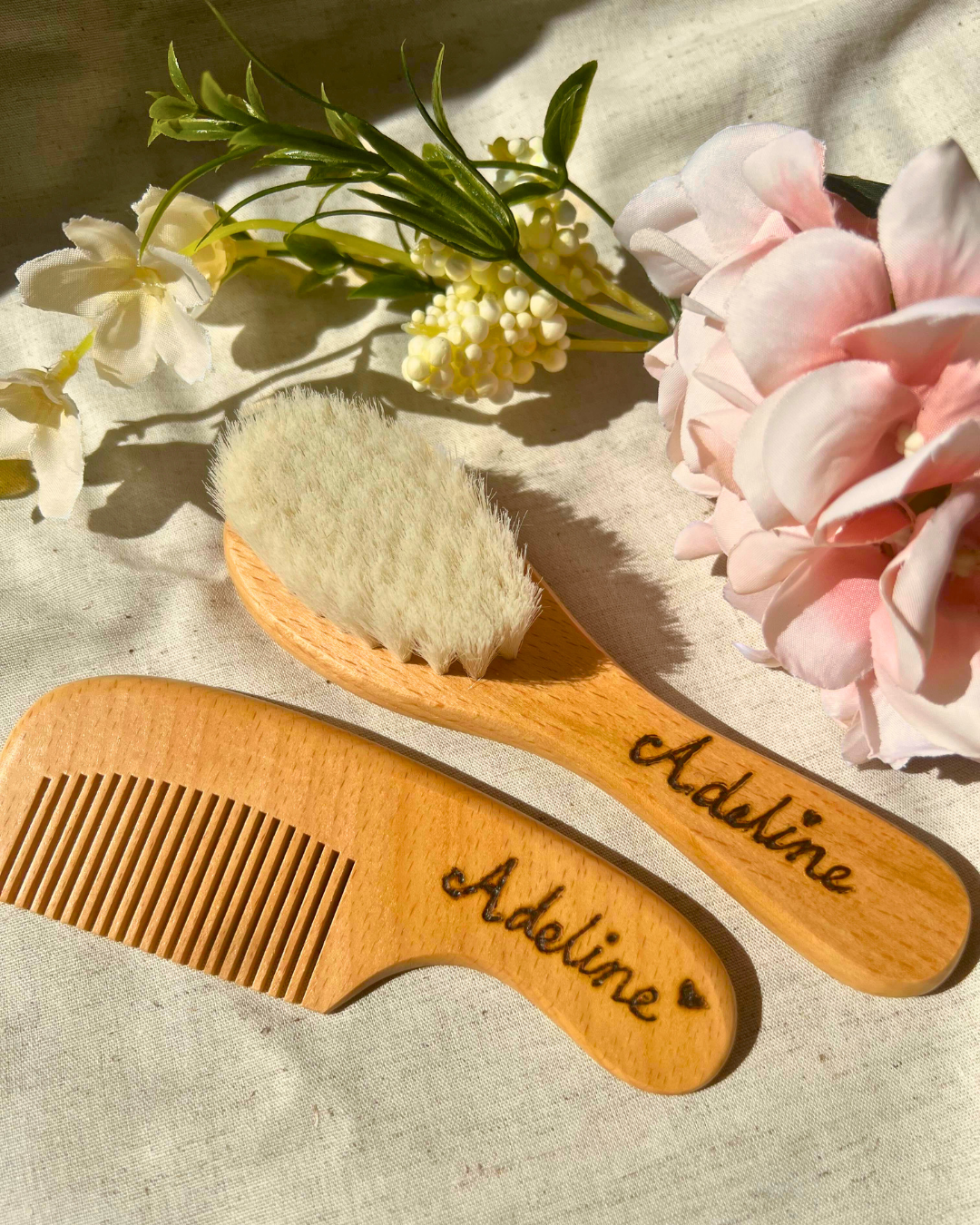 A wooden baby hairbrush and comb set engraved with the name "Adeline," displayed on a soft cream fabric with delicate pink and white flowers in the background. The natural sunlight enhances the gentle textures and warm tones of the scene.